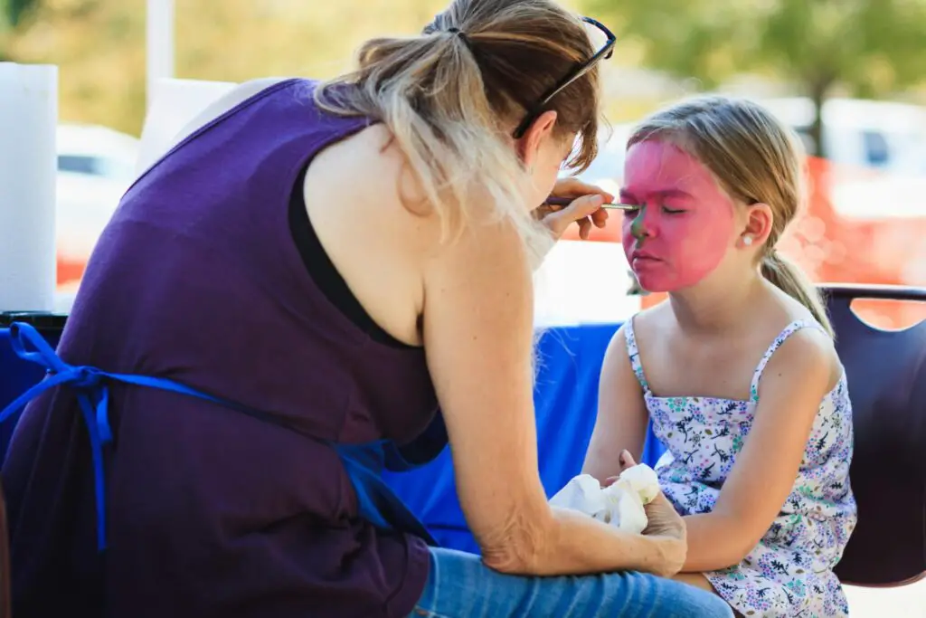 A woman artist skillfully applies vibrant face paint to a girl's face outdoors during a sunny day.